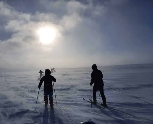 Verschillende mensen skiën in een besneeuwd, winderig landschap met slecht zicht en de zon schijnt door een bewolkte hemel.