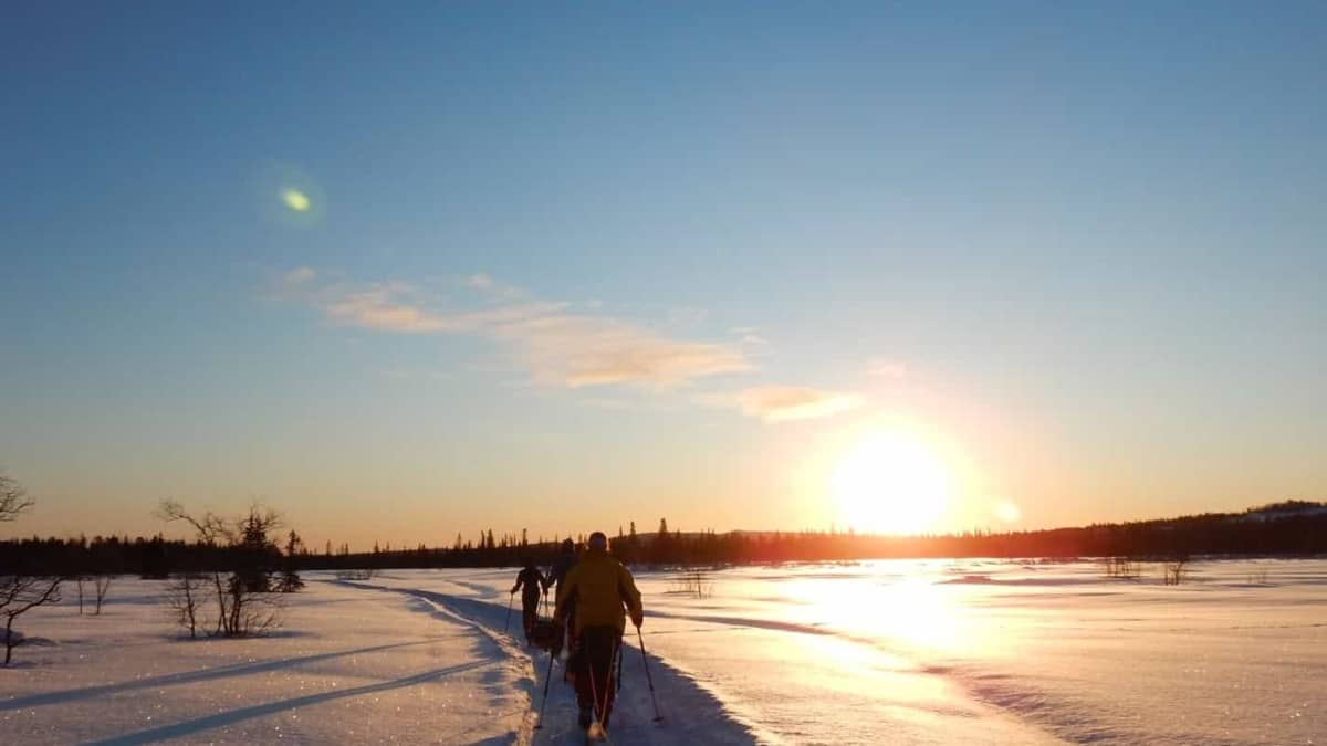Een persoon loopt bij zonsondergang in een besneeuwd landschap en trekt een slee over een pad.
