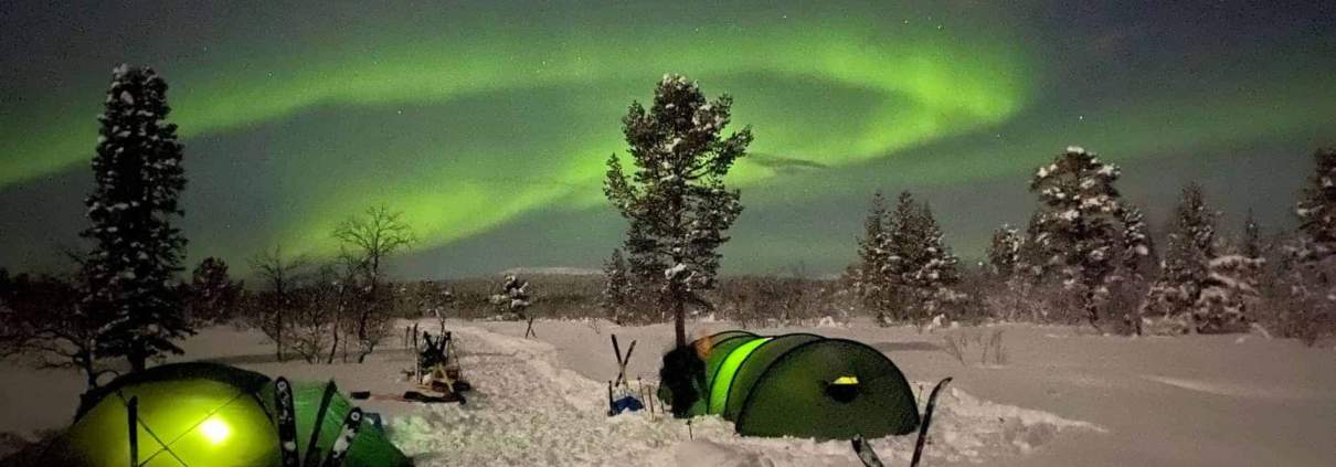 Twee groene tenten opgezet op besneeuwde grond onder de nachtelijke hemel, verlicht door de groene gloed van het noorderlicht, met bomen en skistokken op de achtergrond.