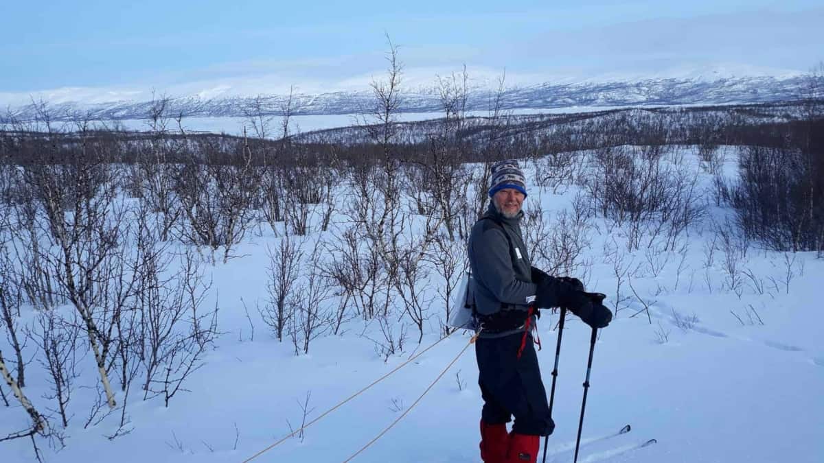 Een persoon in winteruitrusting staat in een besneeuwd landschap met schaarse bomen, houdt skistokken vast en is met een touw aan een slee vastgemaakt. Op de achtergrond zijn met sneeuw bedekte heuvels zichtbaar onder een helderblauwe lucht.