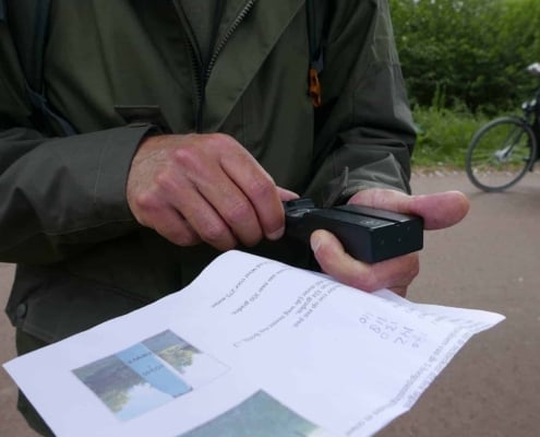 Frank Moison over de Navigeren met kaart en kompas workshop in Nederland 7 Een avontuur in een groene jas staat buiten en navigeert met een gps-apparaat in de hand, terwijl hij een kaart vol afbeeldingen vasthoudt. Op de achtergrond fietst iemand voorbij en je ziet een groen landschap in de verte.