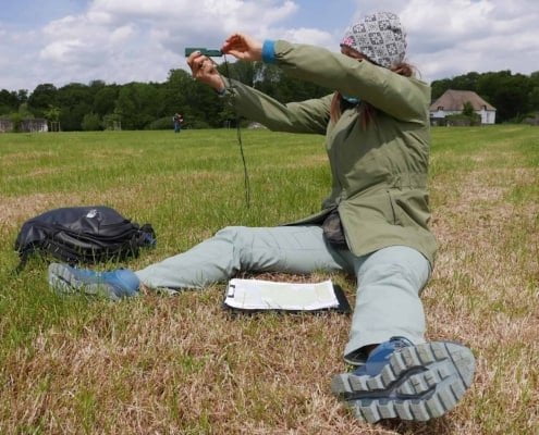 Ready for adventure? Picture this: you're sitting on a green lawn, with a map and backpack at hand. You're wearing a green jacket and light blue pants, perfect for nature. For accurate navigation, you're firmly holding a handy device. In the background, you can see beautiful buildings and tropical trees under a partly cloudy sky. Discover new places and let the adventure begin!     