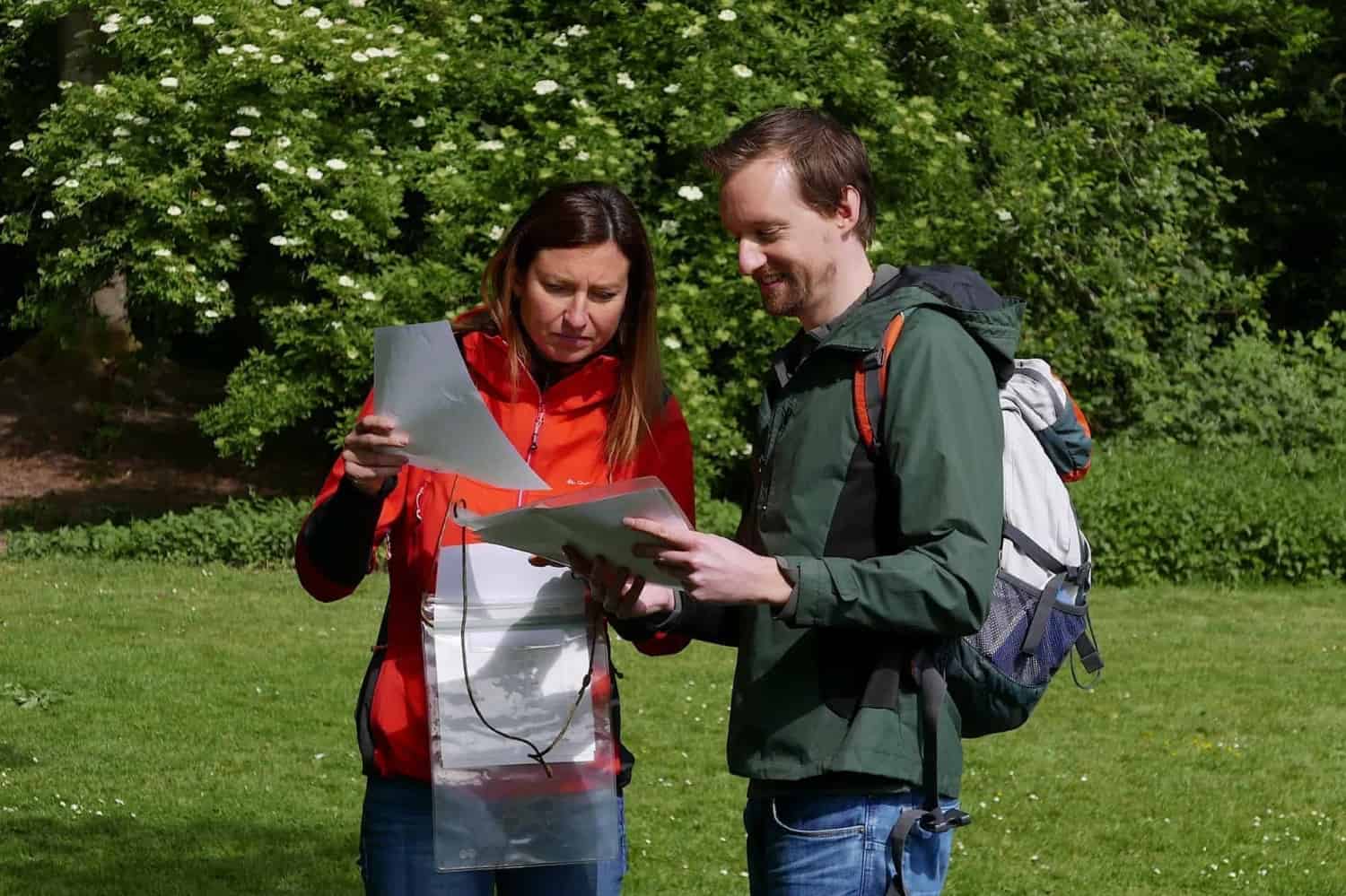 Determining location on a staff map Two adventurers stand outside on a grassy field, surrounded by greenery. Wearing jackets and with a backpack at hand, they attentively study a map. The sun illuminates the scene, perfect for a clear and exciting adventure!