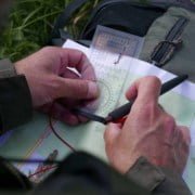 Determining location on the map with a map angle meter Discover the ultimate adventure experience! A person in a green jacket holds a compass and pen over a topographic map. In the background, a green backpack lies on the grass β perfect for map reading and outdoor discoveries. Prepare for your next adventure!