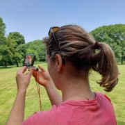 Kaart en kompas navigatie workshop_20 Avontuurlijk bezig in Utrecht: Een persoon met een paardenstaart en roze shirt staat in een groen park. Met een klein spiegeltje en zonnebril op het hoofd, lijkt het ook alsof ze oefenen met kaart en kompas tijdens een spannende workshop. Ontdek Utrecht op soepel wijze!
