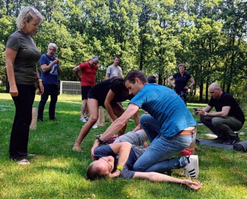 Studio Baak on the Outdoor First Aid Essentials in the Netherlands 5 Een groep mensen traint zelfverdedigingstechnieken in de buitenlucht. Op de voorgrond demonstreert een instructeur een manoeuvre op een cursist die op de grond ligt. Dit lijkt op een Outdoor EHBO-workshop waar volwassenen actief meedoen en toekijken. Omringd door bomen en dicht bij een voetbaldoel, beleef je hier avontuur en leer je essentiële vaardigheden in de frisse buitenlucht.