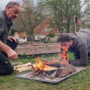 houthak en kampvuur workshop_5 Twee mannen zijn bezig met een klein kampvuur. De een gebruikt zijn jas als windscherm, terwijl de andere het vuur aanwakkert. Omgeven door tuinen en bomen, bij een huis op de achtergrond, voelt het als een spontane houthak- en kampvuurworkshop. Perfect voor avonturiers die van buitenactiviteiten houden!