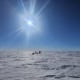 Snowmobiles drive across a vast ice plain under a clear blue sky.