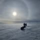 Two people with snowmobiles in the snow under a halo around the sun.