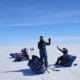 Three people with snowmobiles in a vast white snowy landscape under a clear blue sky.